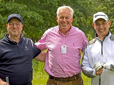 Three men in casual golf attire stand together outdoors, smiling at the camera. Green trees and grass in the background suggest a golf course setting.