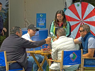 Four people sit outdoors at a table with drinks, in front of a colorful spinning wheel. They appear to be enjoying a casual social event.