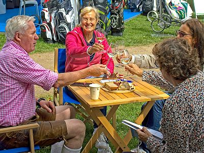 Four people sit around a wooden table outdoors, toasting with wine glasses. They are in casual attire, surrounded by greenery and golf bags.