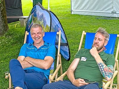Two men sit on deck chairs outdoors; one smiles, the other contemplates, displaying a tattooed arm. A small tent and green grass are in the background.