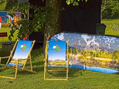Two deck chairs with scenic golf designs on grass, next to a promotional banner, with trees and a tent in the background.