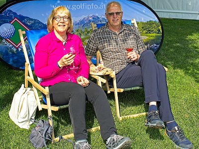 A man and a woman sitting on chairs at an outdoor event, holding drinks. Behind them is a banner with a scenic background, on a sunny day in open grass field.