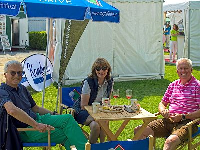 Three people sitting outdoors at a table with parasols, enjoying drinks, indicating a casual and pleasant gathering at an event or fair.