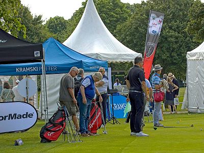 Golf event with people browsing equipment under white tents, flags, and lush greenery. Casual attire, relaxed setting.
