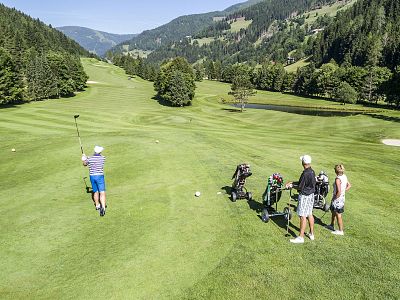 Golfers play on a green course surrounded by mountains and trees under a clear blue sky on a sunny day.