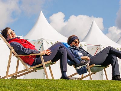 Two individuals relax on deck chairs on green grass, with white tents and a blue sky with clouds in the background.