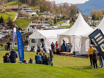 People enjoying an outdoor event with white tents and flags set up on a grassy area, surrounded by a picturesque hillside village under a clear sky.