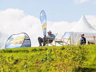 Two individuals sitting on deck chairs near a tent on a grassy hill under a clear sky during a countryside event. Flags and blooming bushes are visible.