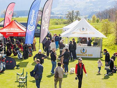 People browse vendor booths at an outdoor event on a sunny day, with colorful banners and green hills in the background.