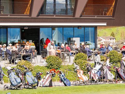 People sitting outside a building with tables and chairs, enjoying meals. Golf bags line the foreground, suggesting a golf event or club scene.
