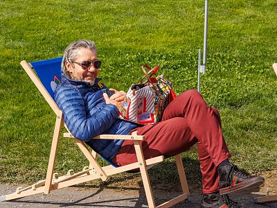 Person sitting in a foldable chair on grass, wearing sunglasses and a blue jacket, enjoying a sunny day.