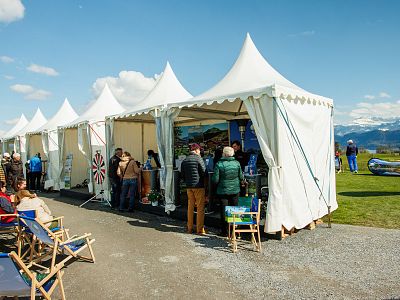 A row of white tents at an outdoor event, people browsing, chairs on the side, and a picturesque mountain view under a blue sky.