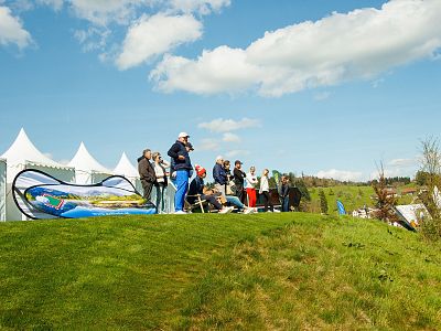 People standing on a grassy hill at an outdoor event with white tents and a blue sky with clouds.