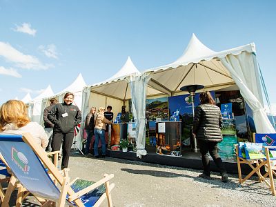 People enjoying an outdoor festival with large white tents, clear sky, and deck chairs. It's a sunny day with people walking and relaxing.