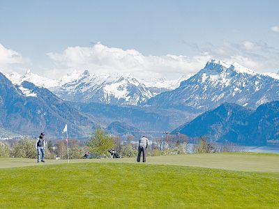 Two golfers on a green golf course with snow-covered mountains in the background under a clear blue sky.