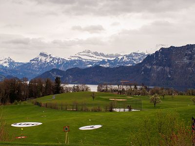 A scenic golf course with lush greenery, set against snow-capped mountains and a cloudy sky, with a lake in the background.