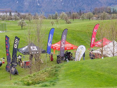 Tents and flags on a golf course, with equipment and branding visible. Green grass, trees, and mountains set a scenic backdrop.