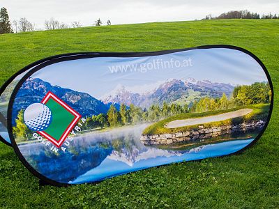 A foldable golf event sign with scenic mountain and lake background, placed on a green field.