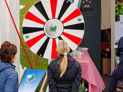 Two people play a spin-the-wheel game with a red and black wheel at an outdoor event, featuring a prize table with a red checkered tablecloth.