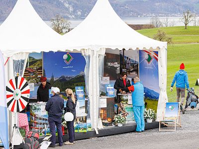 People interacting at a booth with promotional items under white tents. Scenic hills and water are visible in the background.