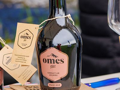 Close-up of a black bottle labeled 'omes 1997', with pink logo, featuring mountain graphic and attached tags, placed on a table with wine glasses nearby.