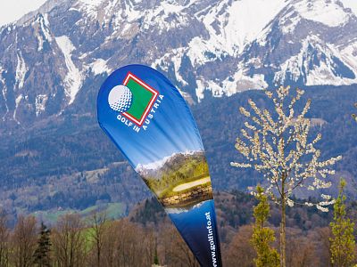 Teardrop-shaped banner promoting golf in Austria, set against snow-covered mountains and blossoming trees under a clear blue sky.