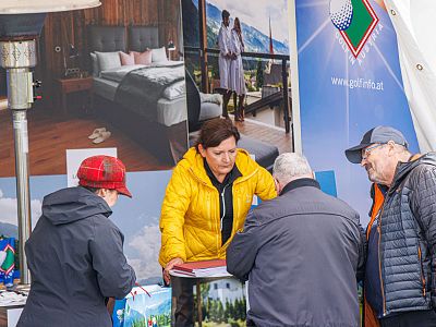 Woman in yellow jacket at a booth talking with three people. Banners with lifestyle images in background.