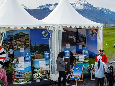 White market tents with colorful displays and people browsing, set against a mountain backdrop on a clear day.