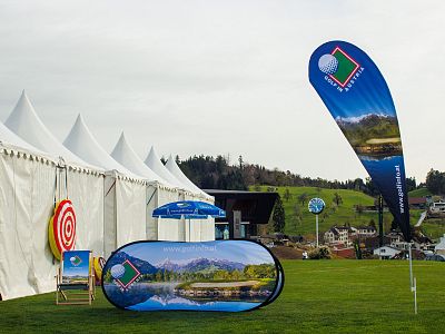 Outdoor event setup with white tents, landscape banners, and a grassy field in a rural area with hills in the background.