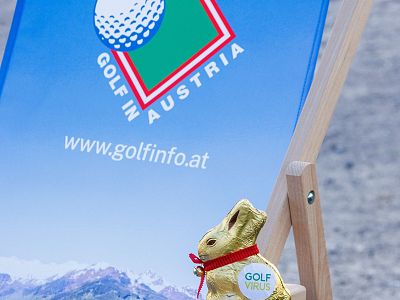 A deckchair with 'Golf in Austria' branding and a chocolate bunny with a golf-themed medallion in front of a snowy mountain background.