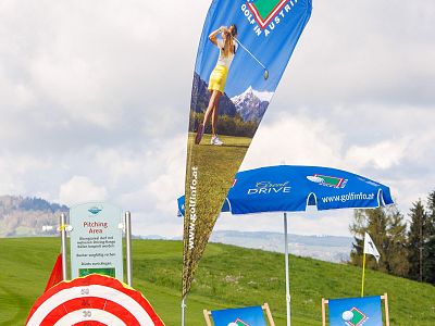 Scene with golf signage, including a flag with a golfer's image, a branded umbrella, and a bullseye target on a grassy field.