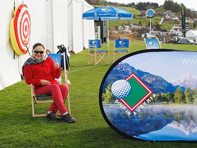 Person in red sitting on a chair next to a golf-themed banner at an outdoor event, with scenic mountains and promotional items in the background.