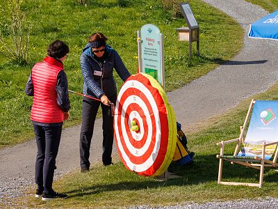Two women at an outdoor archery range, one pointing at a large target. A sunny path and some chairs are in the background.