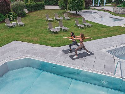 Two people in yoga pose on mats by a poolside, surrounded by green lawn and lounge chairs, under a cloudy sky.