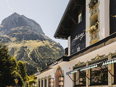 Ein traditionelles Gebäude mit Blumen an den Fenstern vor einem majestätischen Berg unter klarem Himmel, umgeben von grünen Bäumen und strahlendem Sonnenschein.