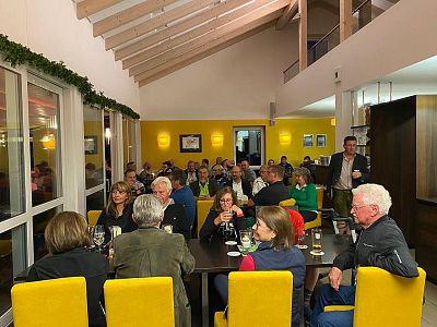 A group of people sitting at tables in a warmly lit room with yellow chairs, engaged in conversation and dining together.
