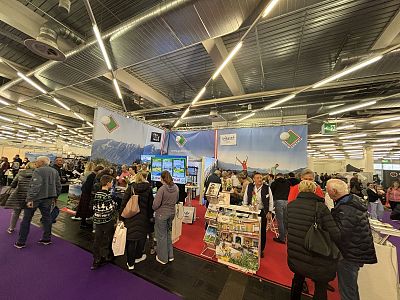 A busy trade show with many people walking around booths displaying products. The ceiling has industrial lighting, and the floor is covered with purple carpet.
