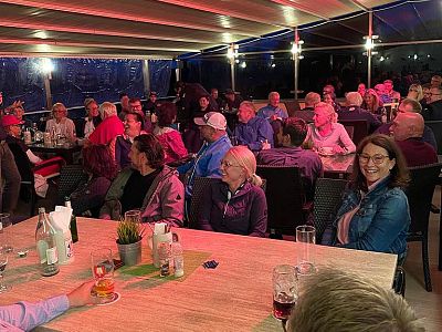 A large group of people seated under a covered outdoor area, socializing and enjoying beverages and snacks at night.