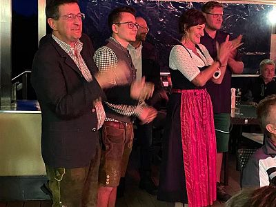 A group of people in traditional Bavarian clothing, including lederhosen and dirndls, clapping and smiling indoors, indicating a festive gathering.