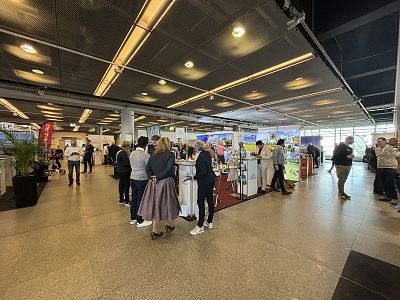 People at a networking event in a modern venue with booths and displays, engaging in discussions. The atmosphere is lively and professional.