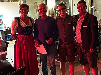 Vier Personen posieren lächelnd in einem Raum. Eine Frau trägt ein Dirndl, die anderen sind leger gekleidet. Im Hintergrund sind Beleuchtung und Dekor zu sehen.