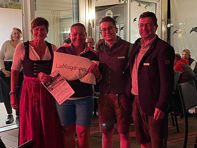 Four people pose for a photo; one holds a certificate and a pillow labeled 'Lieblingsmensch', indoors with people in the background, creating a festive mood.