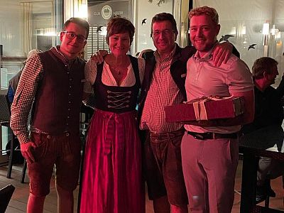 Four people posing together, two in traditional Bavarian outfits, inside a warmly lit room. One person holds a wrapped gift, indicating a festive gathering.