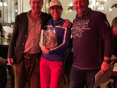 Three people smiling indoors, with the person in the middle holding a gift basket. The atmosphere is casual and warm, with soft lighting.