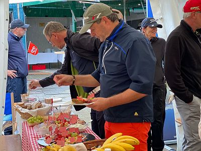 Several people stand at a table with a red checkered cloth, selecting snacks like fruits and pastries at an outdoor gathering.