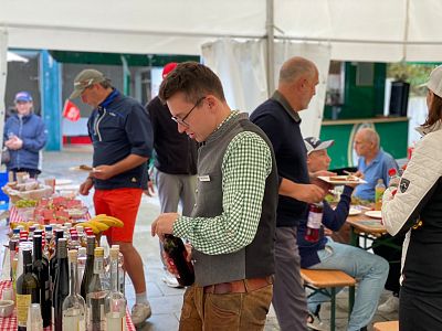 Group of people at a wine tasting event in a tent with tables of wine bottles and snacks. Participants are chatting and sampling drinks.