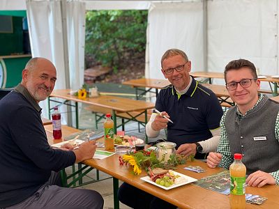 Three men sit smiling at a wooden table under a tent, enjoying a meal together. The table is decorated with flowers and surrounded by greenery, creating a relaxed atmosphere.