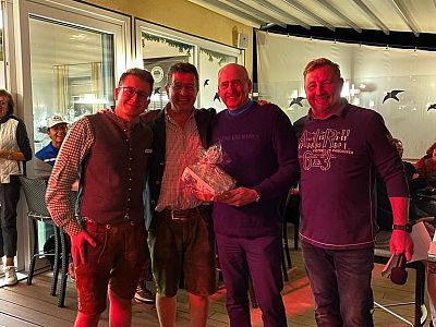 Vier Männer stehen in einem Restaurant oder Café, lächeln und posieren für ein Foto. Einer hält ein Geschenk. Im Hintergrund sind Lichter und Dekorationen sichtbar.