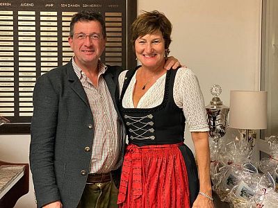 Ein Mann und eine Frau in traditioneller bayrischer Tracht stehen nebeneinander in einem Raum. Sie lächeln in die Kamera. Im Hintergrund sind Geschenke zu sehen.