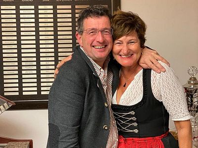 A smiling couple posing together in traditional attire indoors. The man wears a grey jacket, and the woman is in a dirndl. A wall with nameplates is behind them.